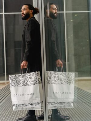 Fashionable man with Debenhams bags reflecting in glass facade in urban setting.