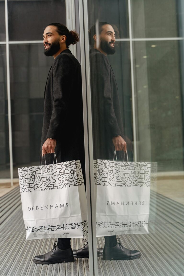 Fashionable man with Debenhams bags reflecting in glass facade in urban setting.